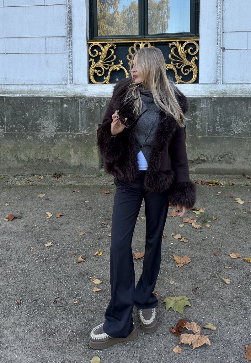 Veste en fausse fourrure marron avec manches texturées, pull en tricot gris, pantalon noir et chaussures marron épaisses avec coutures apparentes.