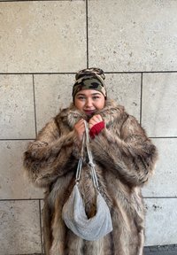 Manteau en fausse fourrure aux tons marron avec un col montant, un bonnet camouflage, et un sac métallique argenté avec une surface texturée et des sangles en chaîne.