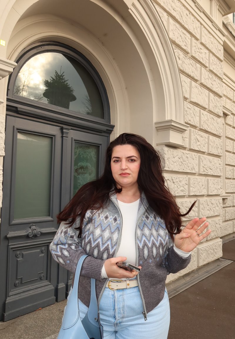 Gestrickter Cardigan mit geometrischen Mustern in Grau und Blau, getragen über einem weißen Hemd; hellblaue Handtasche; steht vor einer dekorativen Tür.