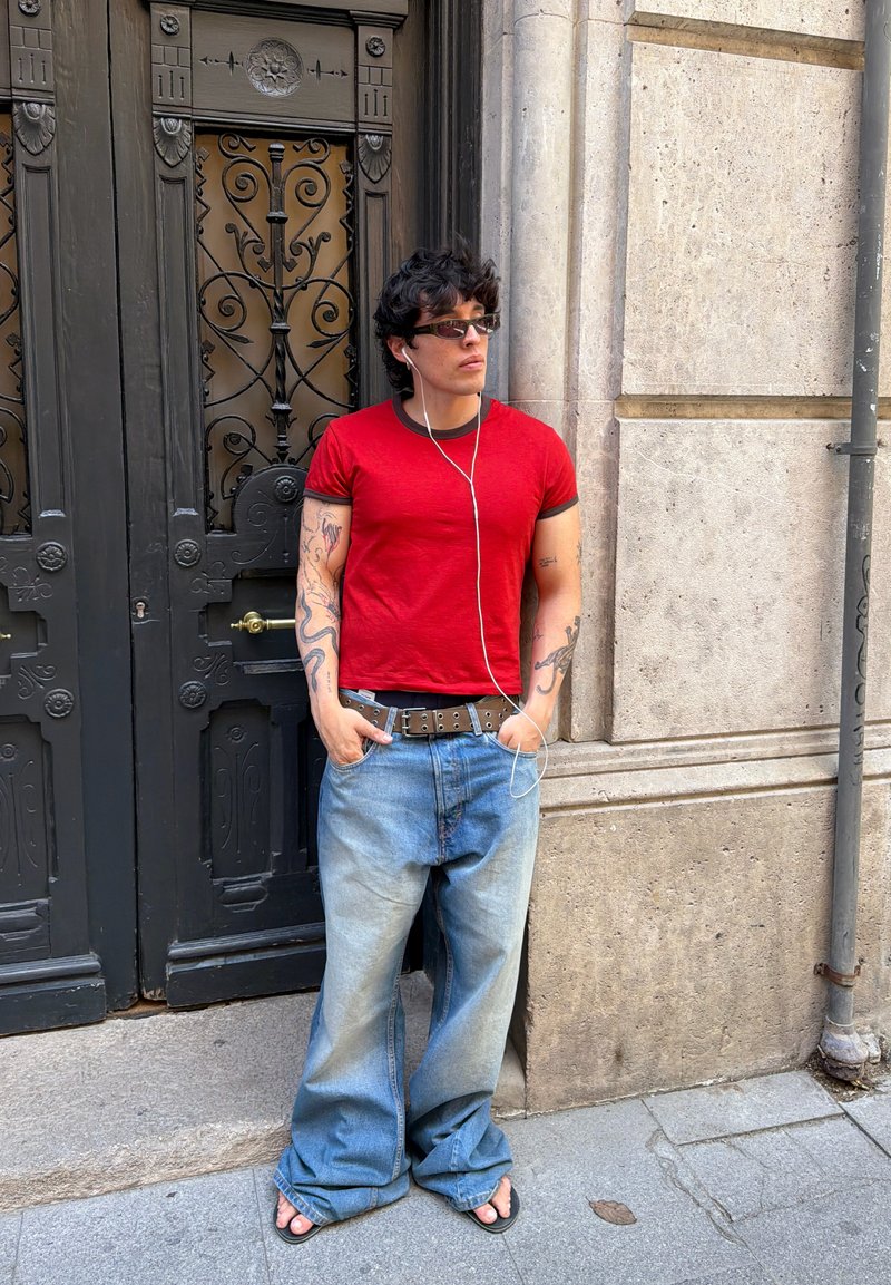 Homme tatoué portant des lunettes de soleil, une chemise rouge, un jean ample et des tongs, debout les mains dans les poches près d'une porte sombre ornée et d'un mur de pierre.