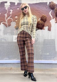 Donna con occhiali da sole neri, maglia a maniche lunghe a quadri gialli, pantaloni a quadri marroni e stivali neri con tacco, in piedi davanti a un muro con arte testurizzata.