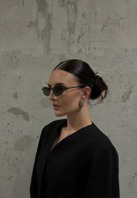 Žena s tmavými vlasy v drdolu, oblečená v černém topu, nosí černé sluneční brýle a stříbrné kruhové náušnice, stojí proti betonové zdi.