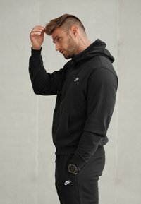 Hanorac negru și pantaloni de trening asortați, confecționați dintr-un material moale. Prezintă un buzunar tip canguru și un logo alb Nike. Textura pare netedă și confortabilă.