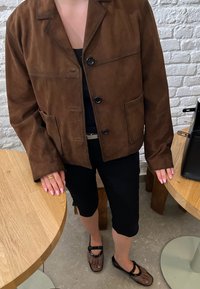 Veste en daim brun, courte, avec un col cranté, quatre poches avant et des boutons noirs. Associée à un short noir et des chaussures en maille.
