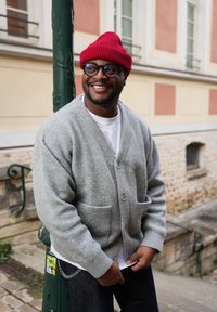 Hombre sonriente con gorro rojo, gafas redondas negras, cárdigan gris sobre camisa blanca, de pie al aire libre cerca de un poste verde y una pared de piedra.