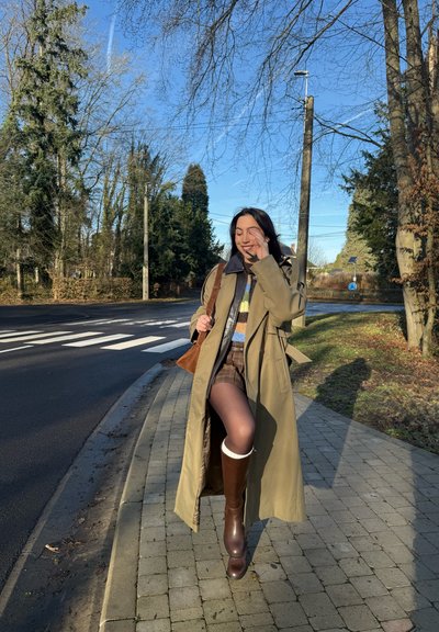 Mujer con abrigo largo beige, botas marrones y bolso marrón caminando por una acera cerca de un paso de peatones en un día soleado.