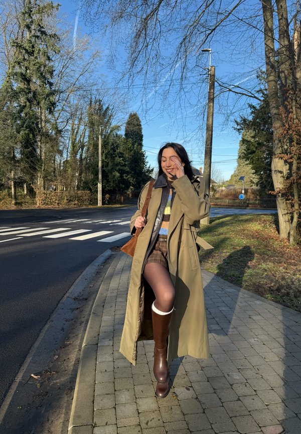 Vrouw in een lange beige jas, bruine laarzen en een bruine tas, lopend op een trottoir vlak bij een zebrapad op een zonnige dag.