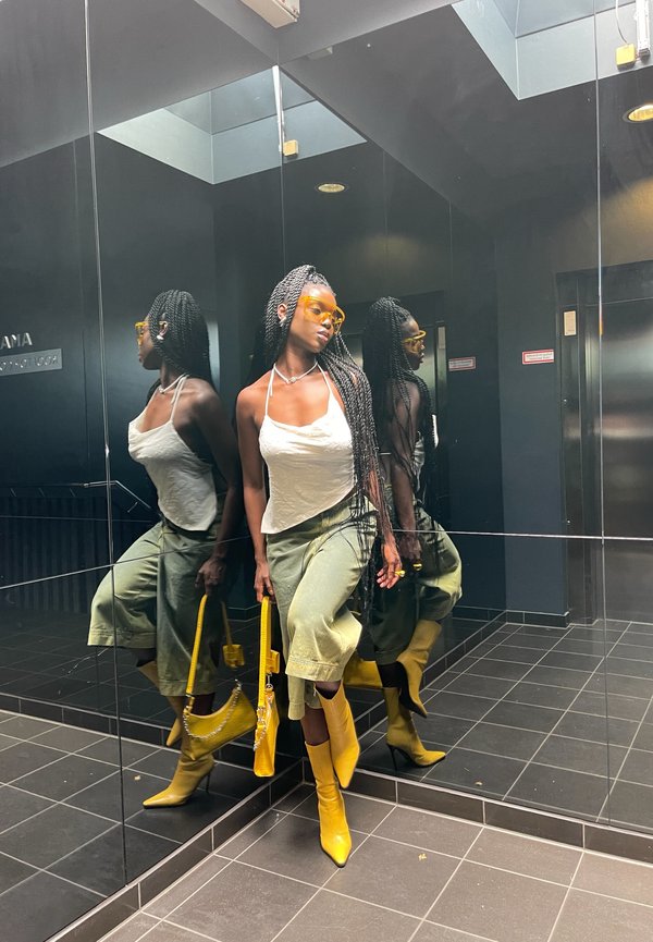 Yellow knee-high boots, olive green wide-leg trousers, and a white top in front of mirrors, showcasing braided hair and yellow sunglasses.