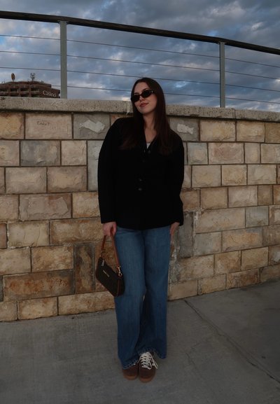 Joven mujer con chaqueta negra, vaqueros azules, gafas de sol, sosteniendo un bolso pequeño, de pie junto a una pared de piedra con barandilla metálica bajo un cielo nublado.