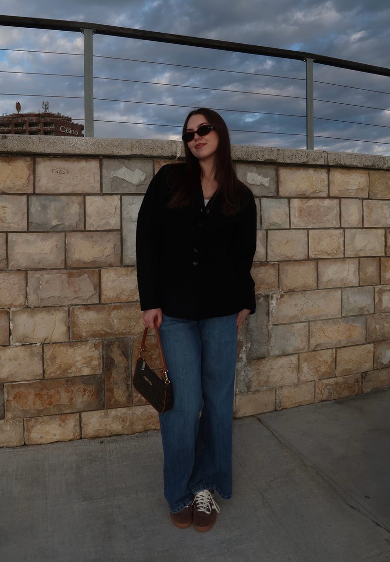 Νεαρή γυναίκα με μαύρο μπουφάν, μπλε τζιν, γυαλιά ηλίου, κρατώντας μια μικρή τσάντα, στέκεται δίπλα σε πέτρινο τοίχο με μεταλλική περίφραξη κάτω από συννεφιασμένο ουρανό.