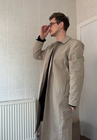 Un homme ajustant ses lunettes, portant un long manteau beige et une chemise noire, se tenant à l'intérieur près d'un mur carrelé blanc et d'un radiateur.