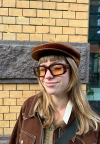 Hnědá kožená čepice, nadměrné brýle s rámem želvovinové barvy a oranžovými skly, a hnědá semišová bunda s bílým límečkem na pozadí cihlové zdi.