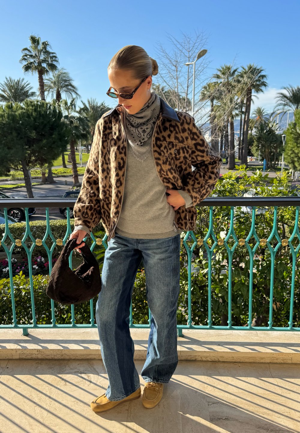 Femme portant une veste à imprimé léopard, un pull gris, un foulard bandana, des lunettes de soleil, un jean bleu et des chaussures beige tenant un sac à main sombre sur un balcon ensoleillé.