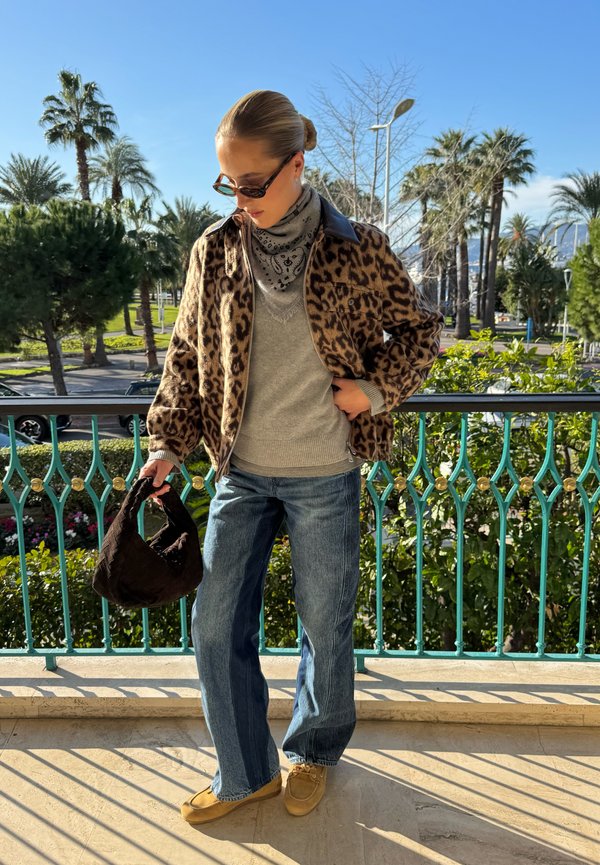 Femeie purtând o jachetă cu imprimeu de leopard, pulover gri, eșarfă bandană, ochelari de soare, blugi albaștri și încălțăminte bej, ținând o geantă închisă la culoare pe un balcon însorit.