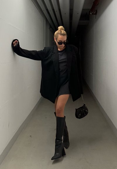 Blazer negro de gran tamaño, vestido negro ajustado, botas de tacón altas negras hasta la rodilla y bolso negro pequeño. Pasillo de color claro con suelo gris.