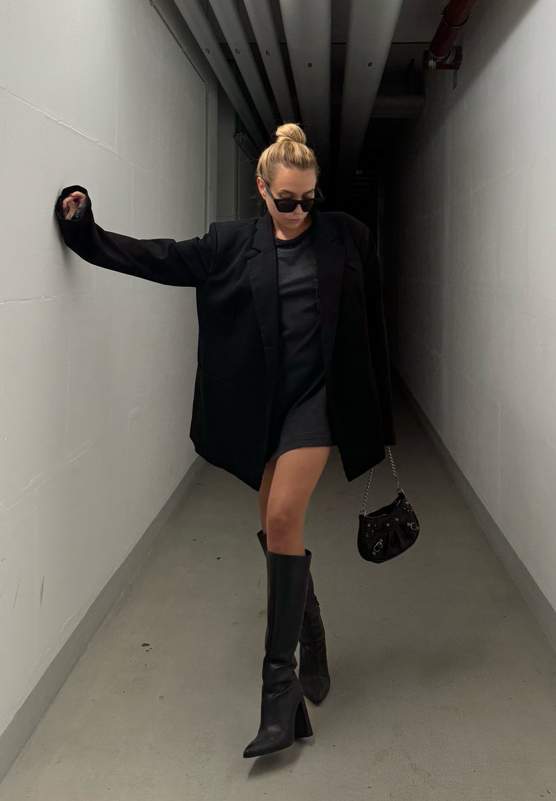 Μαύρο oversized σακάκι, εφαρμοστό μαύρο φόρεμα, μαύρες μπότες με τακούνι μέχρι το γόνατο και μικρή μαύρη τσάντα. Φωτεινός διάδρομος με γκρίζα δάπεδα.
