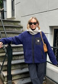 Veste en polaire marine avec fermeture éclair frontale, accents noirs et une poche poitrine. Stylée avec une écharpe gris clair et un sac à main beige.