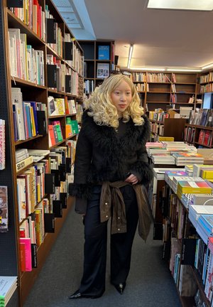 Chaqueta negra de piel sintética con mangas texturizadas, llevada sobre una blusa oscura y un cinturón de cuadros a la cintura, combinada con pantalones negros de pierna ancha y zapatos puntiagudos.