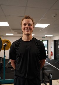 Jauns vīrietis melnā sporta apģērbā stāv un smaidot pozē sporta zālē, fona redzami svarcelšanas aprīkojums un dzeltenās svaru plāksnes.