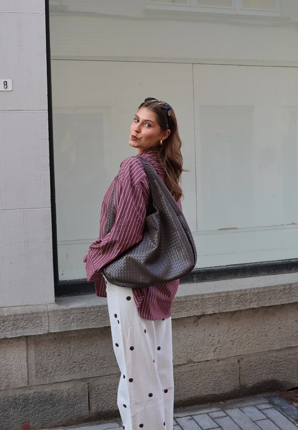 Mujer con cabello largo, usando gafas de sol, camiseta de rayas marrón oscuro, pantalones blancos con puntos negros, llevando un gran bolso gris texturizado al hombro al aire libre.