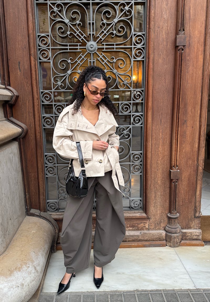 Femme portant une veste beige et un pantalon gris large se tient devant une grille en fer ornée et un cadre en bois, tenant un sac à main noir.