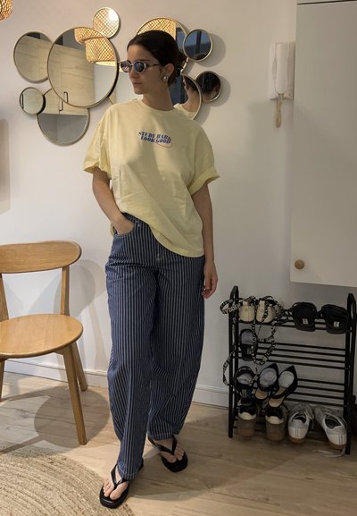 Mujer con gafas de sol, camiseta amarilla y pantalones a rayas, de pie cerca de una silla de madera y un zapatero en una habitación luminosa con espejos redondos en la pared.