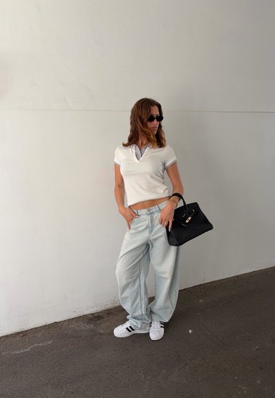 Mujer con polo blanco recortado, vaqueros claros holgados, zapatillas blancas, gafas de sol negras, sosteniendo bolso negro, de pie contra una pared lisa en la acera.