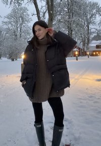 Czarna pikowana kurtka, oversize'owy brązowy sweter z dzianiny, czarne legginsy oraz zielone gumowe buty, na tle zaśnieżonego krajobrazu z drzewami.