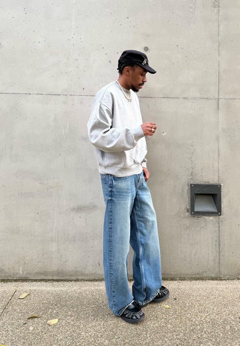 Homme portant un sweat-shirt gris, un jean bleu ample, une casquette noire et des chaussures noires, debout contre un mur en béton, regardant vers le bas.