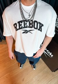Άτομο που φοράει λευκό t-shirt Reebok, πολλαπλές αλυσίδες ασημένιου χρώματος με σταυρό, σκούρο σορτς και μαύρα ασημένια αθλητικά παπούτσια, στέκεται σε ξύλινο πάτωμα.
