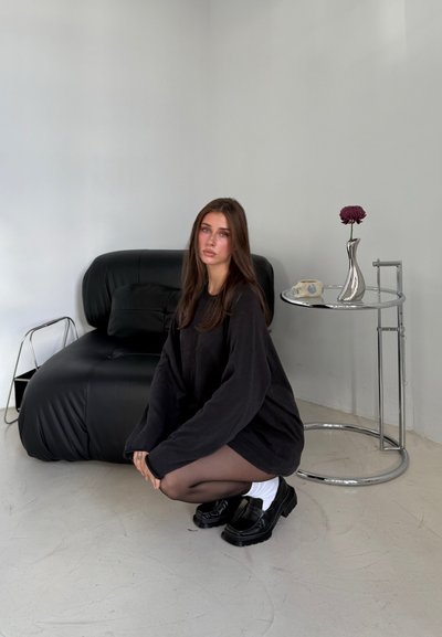 Suéter negro de gran tamaño, medias negras transparentes, zapatos negros gruesos con calcetines blancos. Sofá negro de cuero y mesa auxiliar de cromo con un jarrón.