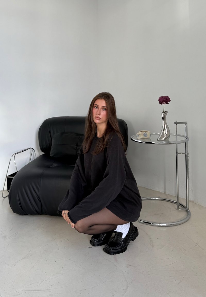 Maglione nero oversize, collant neri trasparenti, scarpe nere massicce con calze bianche. Divano in pelle nera e tavolino in cromo con un vaso.