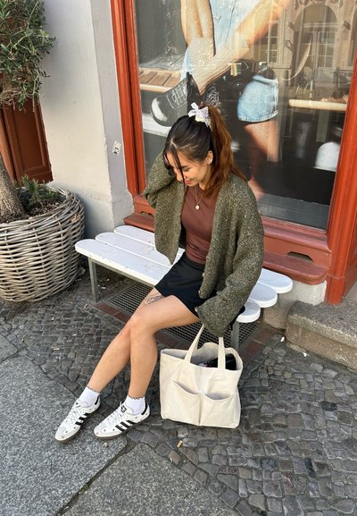 Bolso tote color beige claro con dos bolsillos en el frente, cárdigan verde texturizado, blusa marrón, falda corta negra, zapatillas blancas con detalles en negro.