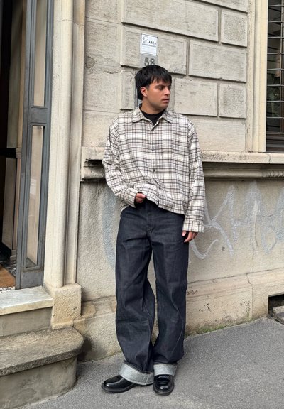 Camisa a cuadros beige y marrón, jeans anchos de pierna oscura con dobladillos remangados y zapatos negros. De pie contra una pared texturizada.