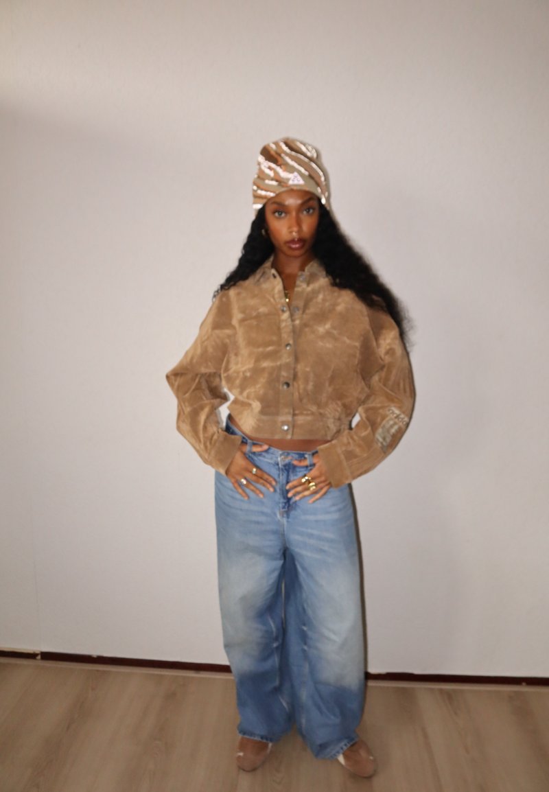 Camisa castanha de veludo cotelê corta, com botões frontais e mangas largas, combinada com jeans azul claro, cintura alta e pernas largas. Lenço para a cabeça com padrão.