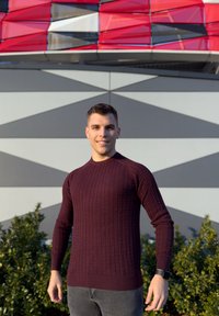 Μπορντό πλεκτό πουλόβερ με υφή καμπίνας, με μακριά μανίκια και ριμπ μανσέτες, συνδυασμένο με γκρι παντελόνι σε γεωμετρικό φόντο.