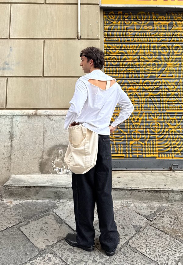 Chemise blanche à manches longues avec des découpes dans le dos, pantalon noir à jambes larges, et un sac en tissu beige. Les textures lisses et les couleurs neutres dominent la tenue.