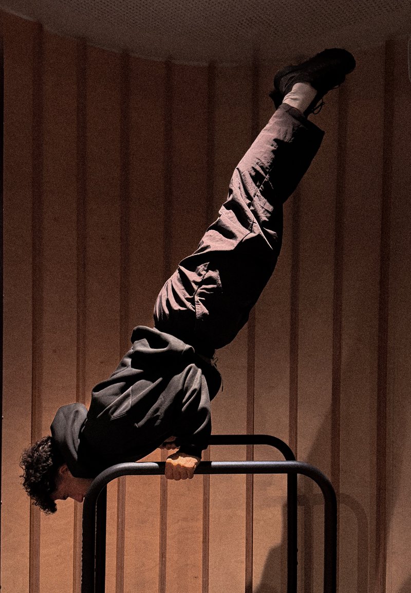 Asmuo dėvintis tamsius drabužius atlieka stovėjimą ant rankų ant lygiagrečiųjų skersinių patalpoje, priešais medinių plokščių sieną.