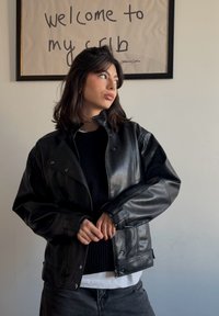 Veste en cuir noir à coupe légèrement oversize, avec un col montant et des détails à boutons-pression, portée avec un pull en maille noir en dessous.