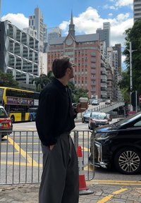Homem de camisa preta e calças cinzentas segura uma chávena de café, de frente para uma paisagem urbana com edifícios modernos e uma estrutura histórica rosa.