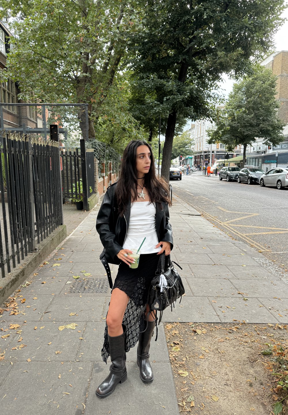 Jaqueta de couro preta, top branco, saia preta de renda com fenda lateral, botas pretas até ao joelho, segurando uma bebida verde, fundo urbano.