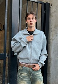 Sudadera gris con cremallera y cuello alto, de corte holgado y puños acanalados, combinada con unos jeans azul desgastados y un cinturón marrón.