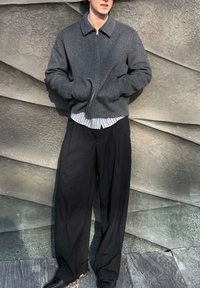 Jeune homme portant une veste grise zippée sur une chemise rayée et un pantalon large noir à fines rayures, debout contre un mur texturé incliné.