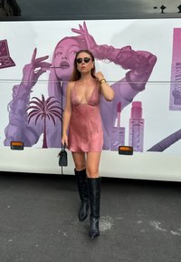 Silk mini kjole i støvete rosa med blonde detaljer, parret med knelengde svarte støvler og en liten svart veske, mot en fargerik bussbakgrunn.