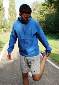 Blå Nike jakke med hætte, grå Nike shorts og grå træningssko. Personen strækker deres ben på en belagt sti.