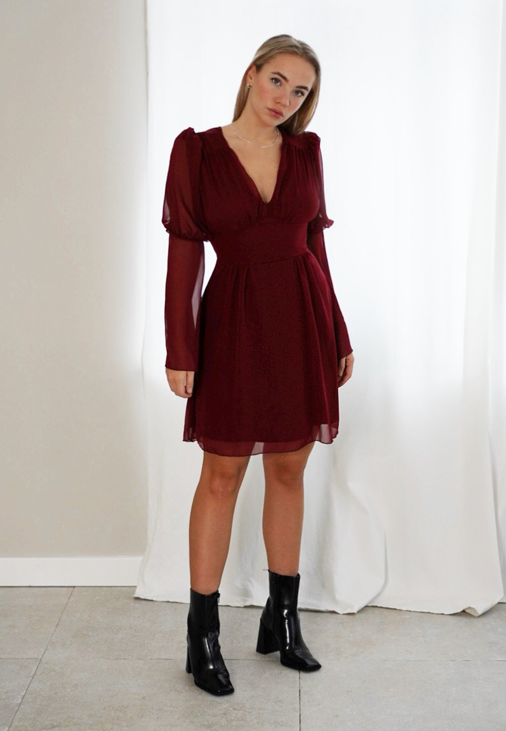 Vestido bordeaux com mangas longas e bufantes, e decote em V, feito de tecido transparente. Combinado com botins pretos de salto.