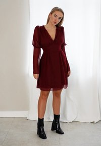 Vestido bordeaux com mangas longas e bufantes, e decote em V, feito de tecido transparente. Combinado com botins pretos de salto.