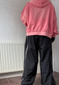 Overmaatse roze hoodie met een onafgewerkt zoom, gecombineerd met zwarte windbreaker broeken met witte zijkanten. Eenvoudig, casual ontwerp.