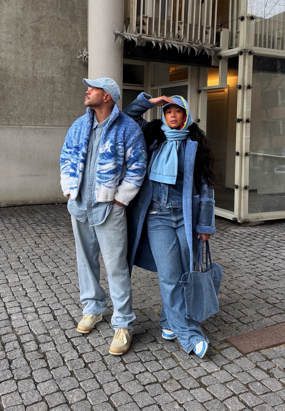 Δύο άτομα στέκονται σε κα cobblestone, ντυμένα με τζιν ρούχα. Ο ένας φοράει ένα μπλε φλις μπουφάν, ενώ ο άλλος έχει ένα μακρύ παλτό και ένα κασκόλ με πολύχρωμες λεπτομέρειες.