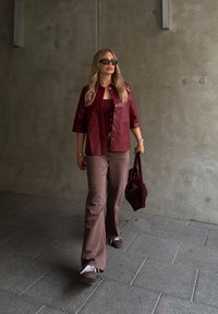 Μαρόνε δερμάτινο πουκάμισο με κουμπιά πάνω από ένα ταιριαστό φανελάκι, συνδυασμένο με καφέ καμπάνα παντελόνι και μια αντίστοιχη τσάντα tote. Φοράει σκούρα γυαλιά ηλίου.